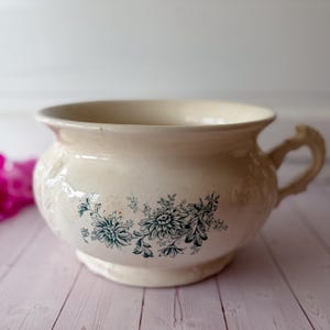 May include: A cream-colored ceramic chamber pot featuring a wide rim, a curved handle, and a decorative transfer print of dark green floral chrysanthemum patterns on the front. The vessel has a rounded body and a raised base with embossed details.