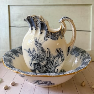 May include: A cream-colored ceramic wash basin and matching pitcher set featuring a dark blue floral iris pattern. Both pieces have scalloped edges with gold trim and intricate blue decorative borders along the rims and the pitcher handle.