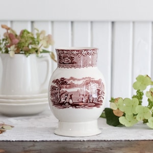 May include: A white ceramic vase with a brown floral pattern. The vase has a round body and a flared base. The pattern features a landscape scene with trees, a lake, and a building.