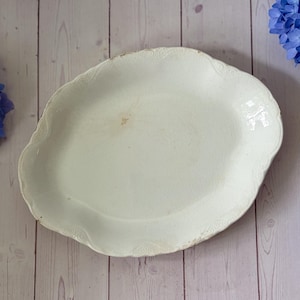 May include: An oval-shaped cream-colored ceramic platter featuring a scalloped edge with embossed detailing. The surface displays a subtle crackle glaze finish and a small brown discoloration mark near the center of the dish.