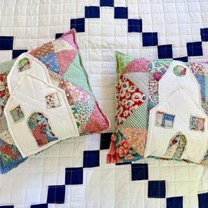 May include: Two square throw pillows featuring a patchwork design made from various floral and patterned fabrics in shades of pink, blue, green, and red. Each pillow has a white fabric applique shaped like a house with windows and a door.