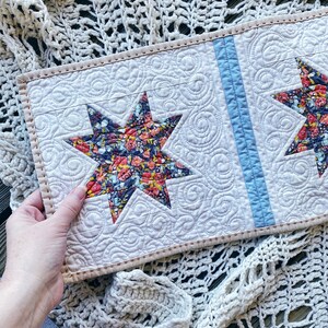 Puede incluir: Un camino de mesa acolchado blanco con una franja azul en el centro. El camino de mesa presenta dos estrellas con estampado floral en cada extremo.