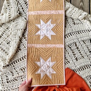 Puede incluir: Un camino de mesa acolchado con un patr&oacute;n geom&eacute;trico de estrellas en tonos de blanco, rosa y beige. El camino de mesa est&aacute; hecho de una tela suave y texturizada y tiene un ribete rosa y blanco.