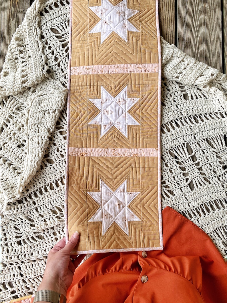 Puede incluir: Un camino de mesa acolchado con un patr&oacute;n geom&eacute;trico de estrellas en tonos de blanco, rosa y beige. El camino de mesa est&aacute; hecho de una tela suave y texturizada y tiene un ribete rosa y blanco.