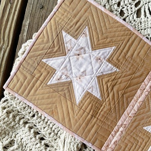 Puede incluir: Un tapiz de pared acolchado con un dise&ntilde;o de estrella en el centro. La estrella est&aacute; hecha de tela blanca con un patr&oacute;n floral y est&aacute; rodeada de una tela marr&oacute;n con un patr&oacute;n de chevron. Los bordes del tapiz de pared est&aacute;n recortados con una tela rosa.