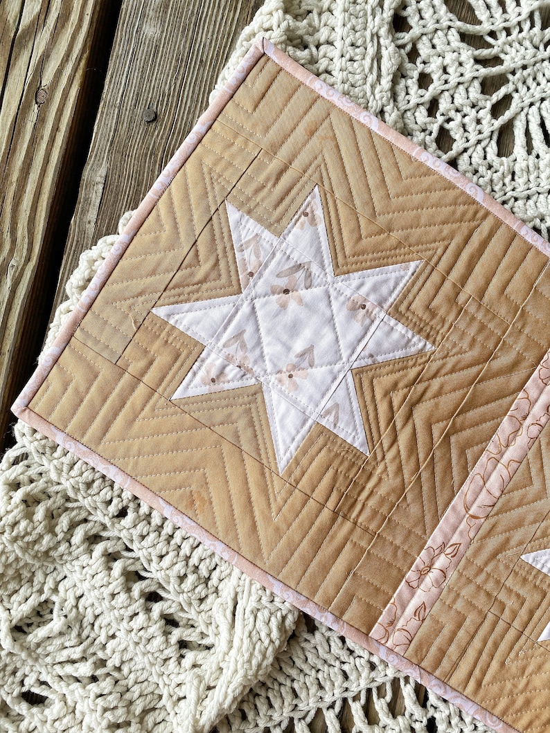 Puede incluir: Un tapiz de pared acolchado con un dise&ntilde;o de estrella en el centro. La estrella est&aacute; hecha de tela blanca con un patr&oacute;n floral y est&aacute; rodeada de una tela marr&oacute;n con un patr&oacute;n de chevron. Los bordes del tapiz de pared est&aacute;n recortados con una tela rosa.