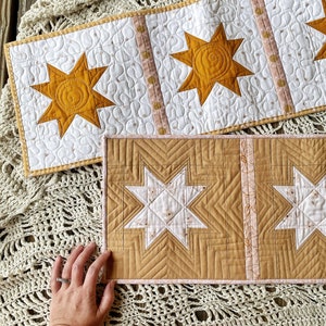 Puede incluir: Dos caminos de mesa acolchados con un dise&ntilde;o de estrella. El camino de mesa superior tiene tres estrellas grandes de color amarillo mostaza. El camino de mesa inferior tiene dos estrellas grandes de color blanco y amarillo mostaza.