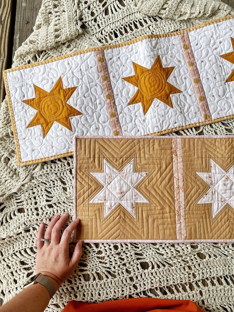 Puede incluir: Dos caminos de mesa acolchados con un dise&ntilde;o de estrella. El camino de mesa superior tiene tres estrellas grandes de color amarillo mostaza. El camino de mesa inferior tiene dos estrellas grandes de color blanco y amarillo mostaza.