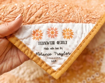 May include: A yellow fabric label with the text "Fernweh Quilt" and "Made with love by Melanie Taylor 2023". The label is attached to a peach-colored quilt with a floral pattern.