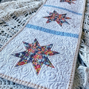Puede incluir: Un camino de mesa blanco con un dise&ntilde;o acolchado que presenta tres estrellas con estampado floral. El camino de mesa tiene una franja azul claro entre cada estrella y un borde cosido marr&oacute;n claro.