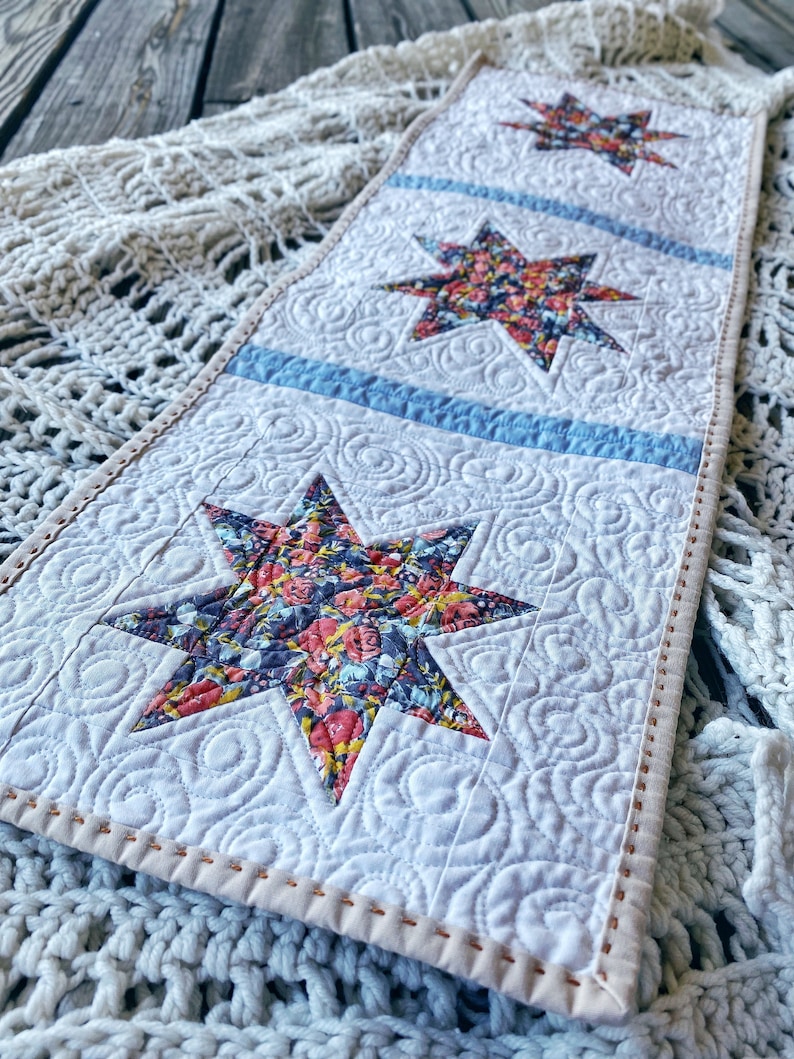 Puede incluir: Un camino de mesa blanco con un dise&ntilde;o acolchado que presenta tres estrellas con estampado floral. El camino de mesa tiene una franja azul claro entre cada estrella y un borde cosido marr&oacute;n claro.