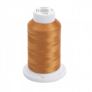 May include: A spool of golden-brown thread, tightly wound around a white plastic base. The thread has a subtle sheen, and the spool's top is also white, with printed text. Ideal for sewing, embroidery, or crafting projects.