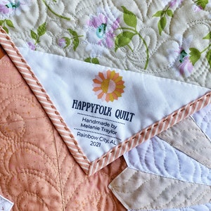 May include: A white label with the text "HAPPY FOLK QUILT" in a sunflower design. The label also includes the text "Handmade by Melanie Traylor, Rainbow City, AL 2021". The label is sewn onto a quilt with a floral pattern.
