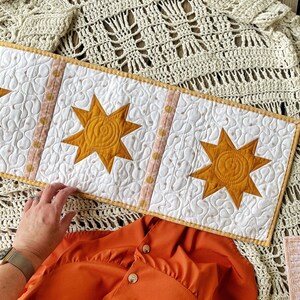 Puede incluir: Un camino de mesa acolchado blanco con un dise&ntilde;o de estrella amarilla. El camino de mesa tiene un ribete de lunares rosa y blanco. El camino de mesa est&aacute; sobre una manta de ganchillo.