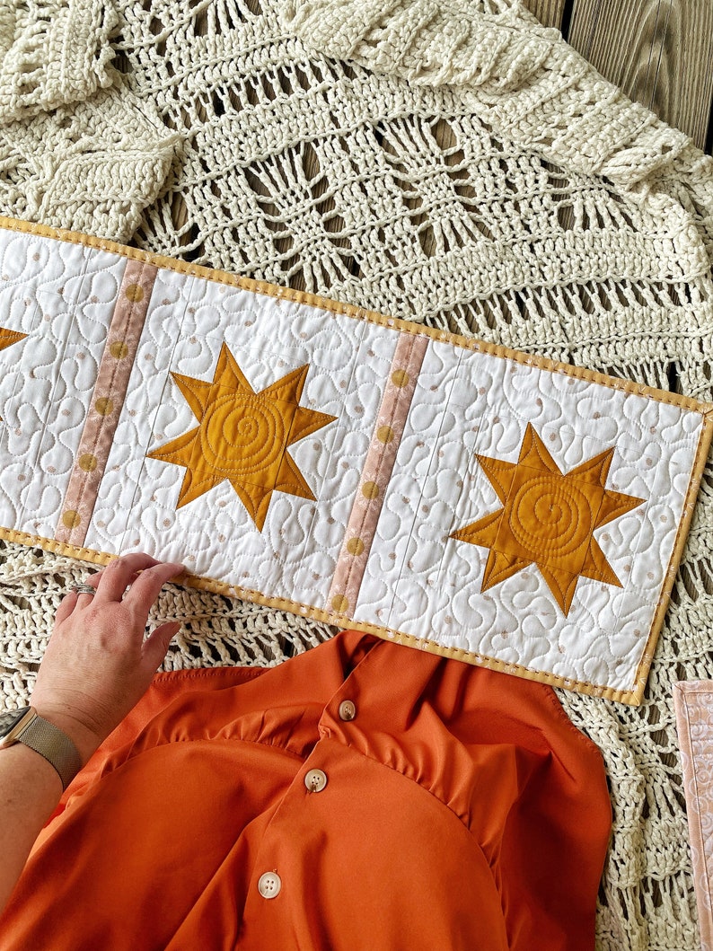 Puede incluir: Un camino de mesa acolchado blanco con un dise&ntilde;o de estrella amarilla. El camino de mesa tiene un ribete de lunares rosa y blanco. El camino de mesa est&aacute; sobre una manta de ganchillo.