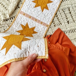 Puede incluir: Un camino de mesa acolchado blanco con un dise&ntilde;o de estrella amarilla. El camino de mesa tiene un borde amarillo y est&aacute; doblado por la mitad. El camino de mesa est&aacute; sobre una manta de ganchillo marr&oacute;n y blanca.