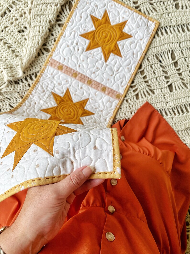 Puede incluir: Un camino de mesa acolchado blanco con un dise&ntilde;o de estrella amarilla. El camino de mesa tiene un borde amarillo y est&aacute; doblado por la mitad. El camino de mesa est&aacute; sobre una manta de ganchillo marr&oacute;n y blanca.