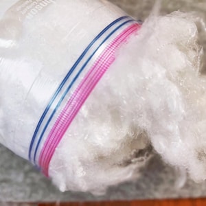 May include: A clear plastic bag filled with fluffy white fiber. The bag is sealed with a pink and blue striped zipper. The fiber is spilling out of the bag.