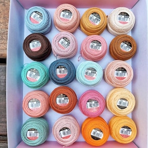 May include: A collection of DMC cotton perle thread balls in a pink box. The thread colors include shades of blue, pink, orange, yellow, brown, and green. Each ball has a label with the DMC logo and a number.