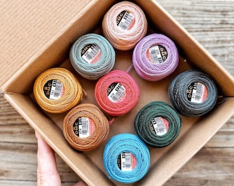 May include: A box of eight spools of DMC embroidery floss in various colors, including pink, purple, teal, yellow, red, green, brown, and blue. Each spool has a white label with the DMC logo and a number.