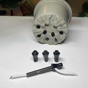 Może przedstawiać: Zestaw trzech czarnych plastikowych nóżek do doniczek i szarej plastikowej doniczki z otworami drenażowymi. Biały sznurek jest przymocowany do czarnego plastikowego kolca do podlewania.