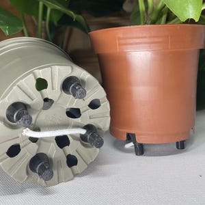 Könnte beinhalten: Zwei Plastik-Blumentöpfe, einer braun und einer grau. Der graue Topf hat Abflusslöcher und eine weiße Schnur um den Boden. Beide Töpfe haben kleine schwarze Füße.