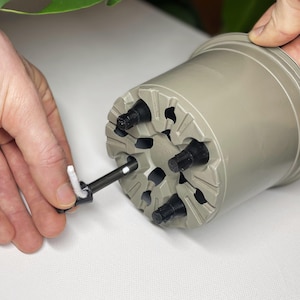 Könnte beinhalten: Ein grauer Plastiktopf mit eingebautem Selbstbewässerungssystem. Der Topf hat fünf schwarze Ausläufe und einen weißen Docht, der in einen der Ausläufe gesteckt ist.