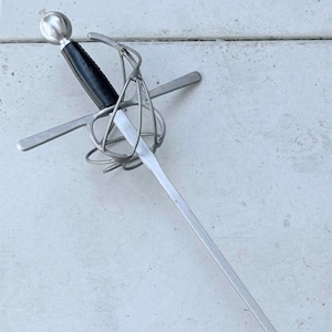 May include: A rapier sword with a long, slender, silver blade. The hilt features a black grip, a complex wirework basket, and a crossguard. The pommel is a rounded silver sphere. The sword is displayed against a white background.