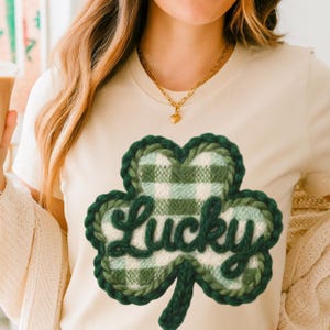 May include: A cream-coloured t-shirt with a green and white plaid shamrock design and the word "Lucky" in cursive. The shamrock is outlined in a darker green. A gold necklace with a heart pendant is visible.