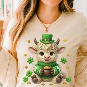 以下が含まれることがあります： クリーム色のTシャツ。緑の帽子とセーターを着た漫画の羊が金のポットを持っています。デザインにはクローバーと金貨が含まれており、聖パトリックの祝日を祝っています。