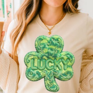 Puede incluir: Camiseta color crema con un gran diseño de trébol texturizado en tonos verdes. La palabra "LUCKY" se muestra en verde. Esta camiseta informal es adecuada para el uso diario.