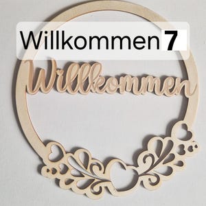 Könnte beinhalten: Ein hölzernes Willkommensschild mit dem Wort "Willkommen" in Schreibschrift, eingebettet in einen kreisförmigen Rahmen. Der Rahmen weist dekorative Ausschnitte von Herzen und Wirbeln auf. Die Zahl "7" steht über dem Wort "Willkommen".