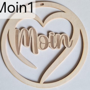 May include: A wooden heart-shaped decoration with the word "Moin" cut out in the centre. The ornament is made of light-coloured wood and has a small hole at the top for hanging. The text "Moin1" is in the upper left corner.
