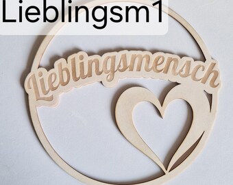 May include: A wooden decorative piece with the German word "Lieblingsmensch" (favorite person) in a cursive font, inside a circle. A heart shape is cut out below the text. The text "Lieblingsm1" is in the upper left corner.