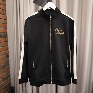 Può includere: Giacca da tuta nera con strisce bianche sulle maniche. La giacca ha un collo alto, una cerniera a tutta lunghezza e due tasche con cerniera. Il testo "Palm Angels" è ricamato in oro sul petto.