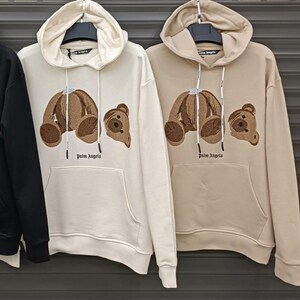 May include: Two cream-colored hoodies with brown embroidered teddy bears on the front. The hoodies have a drawstring hood and a kangaroo pocket. The text "Palm Angels" is printed in black below the bears.