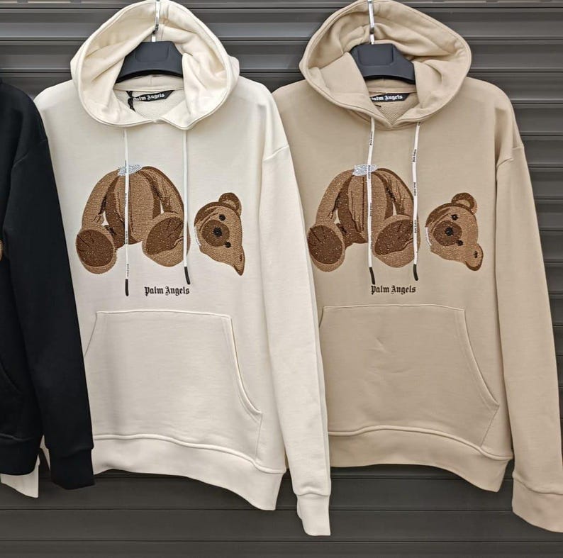 May include: Two cream-colored hoodies with brown embroidered teddy bears on the front. The hoodies have a drawstring hood and a kangaroo pocket. The text "Palm Angels" is printed in black below the bears.