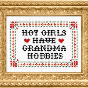 K&ouml;nnte beinhalten: Kreuzstich-Kunstwerk in einem goldenen, verzierten Rahmen. Der wei&szlig;e Stoff zeigt den Text "HOT GIRLS HAVE GRANDMA HOBBIES" in Schwarz, mit roten Herzakzenten. Der Rand hat ein dekoratives Muster in Rot, Gr&uuml;n und Orange.
