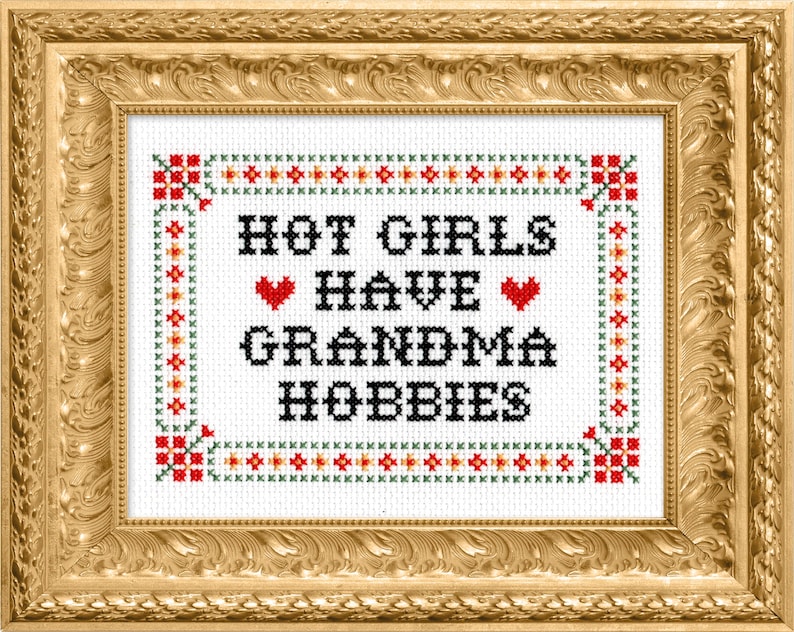 K&ouml;nnte beinhalten: Kreuzstich-Kunstwerk in einem goldenen, verzierten Rahmen. Der wei&szlig;e Stoff zeigt den Text "HOT GIRLS HAVE GRANDMA HOBBIES" in Schwarz, mit roten Herzakzenten. Der Rand hat ein dekoratives Muster in Rot, Gr&uuml;n und Orange.