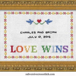 May include: A white cross-stitch embroidery in a gold frame with the text "Charles and Brian July 12, 2015 Love Wins" in colorful thread.