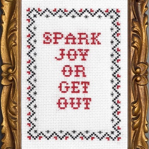以下が含まれることがあります： 白い背景に赤い文字で「Spark Joy or Get Out」と書かれた、額縁に入ったクロスステッチ。フレームは金色の装飾が施されている。