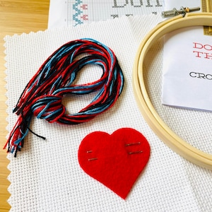 K&ouml;nnte beinhalten: Ein Kreuzstich-Set mit einem h&ouml;lzernen Stickrahmen, einem roten Filzherz und einem Strang rotem, schwarzem und blauem Garn. Das Set enth&auml;lt wei&szlig;en Aida-Stoff mit einem Kreuzstichmuster und dem Text "Don't".