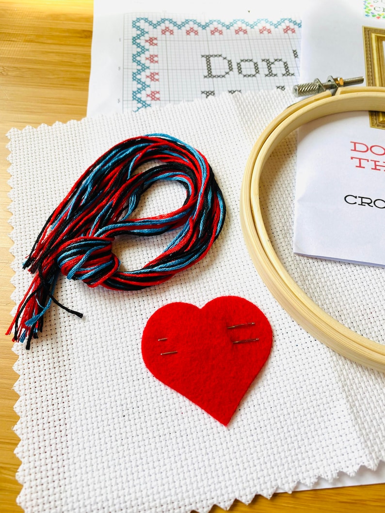 K&ouml;nnte beinhalten: Ein Kreuzstich-Set mit einem h&ouml;lzernen Stickrahmen, einem roten Filzherz und einem Strang rotem, schwarzem und blauem Garn. Das Set enth&auml;lt wei&szlig;en Aida-Stoff mit einem Kreuzstichmuster und dem Text "Don't".