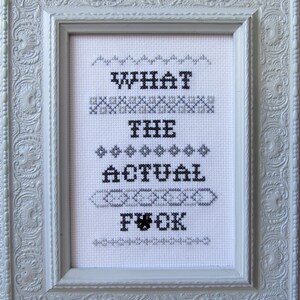 May include: White framed cross-stitch embroidery with the phrase "WHAT THE ACTUAL F*CK" in black thread.