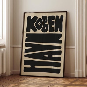 Pode incluir: Um pôster preto e branco com o texto "KOBENHAVN" em uma fonte em negrito e estilizada. O pôster está emoldurado e pendurado em uma parede de um quarto com paredes brancas e piso de madeira.
