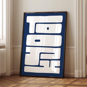 May include: A framed art print featuring a bold, abstract design. The artwork has a dark blue background with large, white, geometric shapes. The print is displayed on a wooden floor, leaning against a white wall. The frame is dark brown.