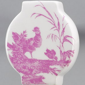 May include: A white ceramic knob with a pink floral design featuring a bird and a plant. The knob is shaped like a rounded rectangle with a small opening at the top.