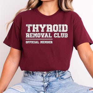 May include: A maroon short-sleeved t-shirt featuring white distressed text that reads THYROID REMOVAL CLUB and OFFICIAL MEMBER. The shirt has a crew neckline and a standard fit, displaying a graphic design centered on the chest area.