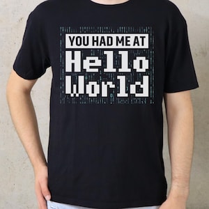 Pode incluir: Camiseta preta com a frase "YOU HAD ME AT HELLO WORLD" em uma fonte pixelizada. A camiseta é de gola redonda. O logotipo The Print Studio está no canto superior esquerdo, e a palavra "BLACK" em um círculo no canto superior direito.