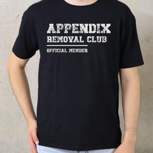 May include: A black short-sleeved t-shirt featuring white distressed-style text on the chest that reads "APPENDIX REMOVAL CLUB" followed by a horizontal line and the words "OFFICIAL MEMBER" in a smaller, clean white font.